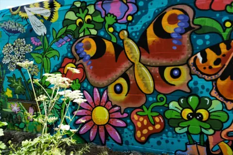 Nick Ballard A painted mural featuring a large yellow, red and orange butterfly surrounded by green plants, pink flowers and red strawberries