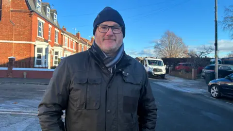 A man wearing a black coat, scarf , glasses and blue hat is stood in front of an icy road. Behind him there's a row of red brick terraced houses and a white truck. The sky is blue and it is a sunny, bright day. 