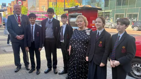 Four boys and a girl from Belfast Royal Academy, in dark school uniform, in front of the new red postbox featuring the yellow cypher of King Charles III. To their left is a tall man in a grey suit with a red tie and a gold chain of office around his neck.
Also in the picture, third from the right, is a blonde-haired woman in a navy and white polka dot dress.