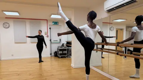 Jaydon, a black teenager, practices an arabesque at a studio barre, lifting one leg high behind them while holding the barre for balance. Dance teacher Grace stands across the room demonstrating the same arm position, with the mirrored wall reflecting the scene in the bright wooden-floored studio.