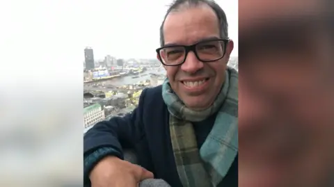 Andrew Wareham Andrew Wareham smiling at the camera in glasses and tartan scarf on a winters day from a balcony looking over a city