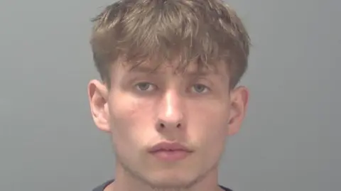 Suffolk Constabulary Jake McMillan is looking at the camera in a police custody photo. He has short light brown hair and blue or green eyes. He has a tufty beard.