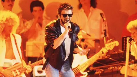 Murray Sanders George Michael singing at Live Aid in a leather jacket and sunglasses, with the band behind him under amber stage lighting.
