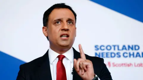Anas Sarwar, who has short black hair, speaks with his left index finger raised. He in front of blue and white Scottish Labour branding. 