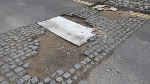 Facundo Arrizabalaga A damaged cobblestone speed bump where some stones are missing, exposing the dirt beneath. A wooden board partially covers the damaged area, and double yellow lines run along the road.