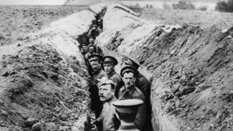 Getty A black and white photo shows a long, narrow trench carved into a field with soldiers stood in it wearing military caps and heavy coats.