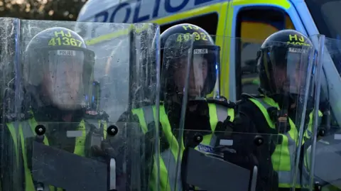 Police officers wearing helmets and high‑visibility vests stand close together holding clear riot shields, with a marked police van parked behind them.