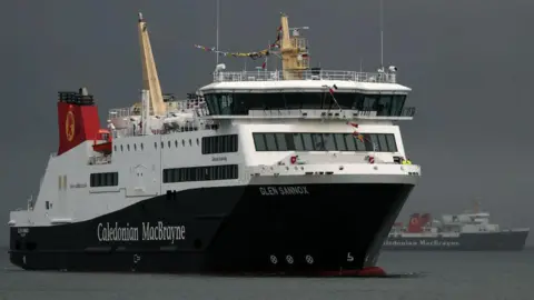 The MV Glen Sannox, a car ferry in characteristic Cal Mac black, white and red livery, is moving port to starboard on grey sea against a deep, dark grey sky. Another ferry can be seen in the background. 