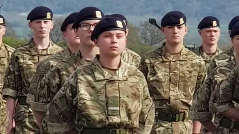 Family Handout Gunner Beck wearing her camouflage military uniform and black cap. She is standing among her fellow soldiers with her arms clasped behind her back and a stern facial expression.