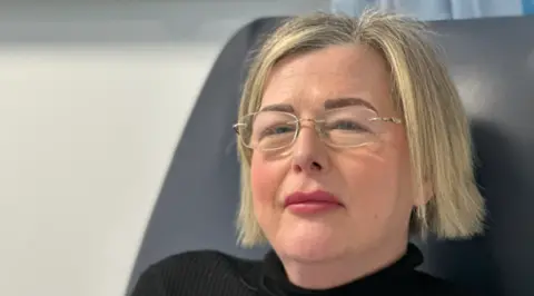 A woman with blonde hair and glasses is looking at the camera. She is sitting down on the right of the photo and wearing a black top.