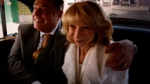 Character Jesse Chadwick dressed in a wedding suit hugs smiling wife Gail in the back of a car.