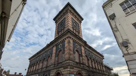 Huw Rees An opulent red and cream bricked building, with a square clock tower in the middle. The clocks have been removed and the large arched windows have been boarded up