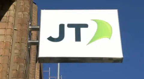 BBC A brick wall to the left holds a metal sign on which is written JT on a white background with a green insignia, the sky in the background is blue with no clouds. 
