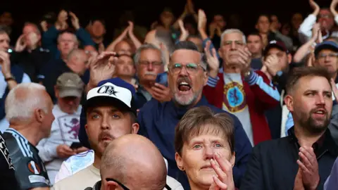 Aston Villa fans cheering in a stadium. They are wearing claret, blue and sky blue hates, jumpers and scarfs colours of the club
