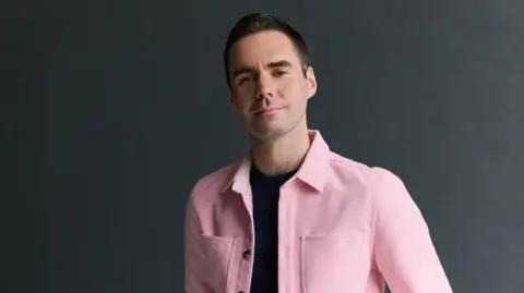 Rich Lakos Joshua McTaggart - a man in a pink jacket with a dark blue t-shirt on.