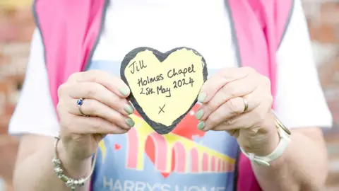 HolmesChapelPartnership  Woman holds stone heart up with the words 'Jill, Holmes Chapel, 25 May' written on it.