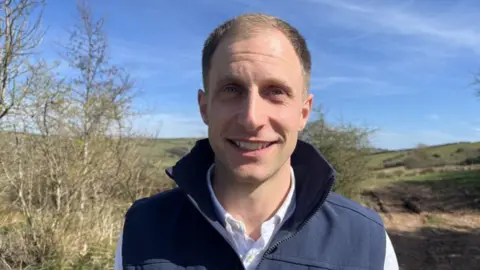 A man with short blonde hair in a white shirt and blue jacket standing in a field with grass behind him