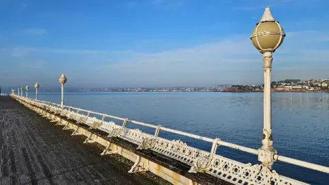 A sunny day picture showing a Victorian pier with cast iron seating and lighting and the sea in the background