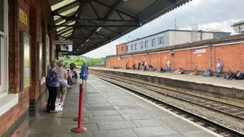 A picture of a railway track taken from a train platform. The platform walls are made from red brick and a few older women are pictured on the platform from behind. There are two small red bollards at the front of the image. 