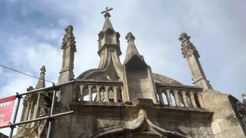 The Poultry Cross. It is a stone, intricately carved structure and there is scaffolding to the left of the building.