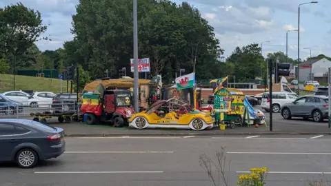 A bright yellow car, caravan and other brightly-coloured pieces of equipment on an island off a busy main road