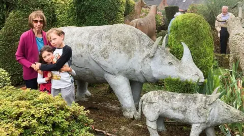 Clare Stephens An older woman and two young children stand in front of two cement rhinos