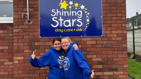 supplied Two woman in blue hoodies smile and hug as they hold their thumbs up to the camera