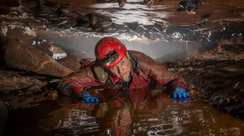 Bartek Biela A caver from the exploratory team wearing waterproofs, blue gloves and a red helmet with a light on on his front between a small gap of rock that sits above and below him. He has his hands on the wet ground in front of him and is looking at the camera.