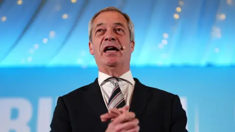 PA Media Nigel Farage giving a speech at a rally in North West Essex on Friday night.  he is wearing a dark suit, white shirt and a tie with thick symmetrical stripes which are blue and silver.  The backdrop is in soft focus and the light blue colour of his Reform UK party. 