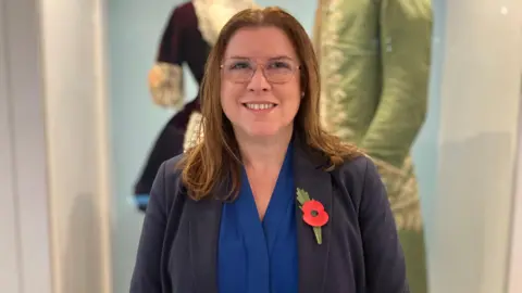 A woman with light brown hair wearing glasses, a blue top and a dark jacket with a red poppy pinned to it, stands in front of a display of old-fashioned clothes while smiling.