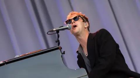 Kieran Frost/Getty Images Tom Odell performing onstage with sunglasses and a black blazer on. He is singing into a microphone and playing a piano. 