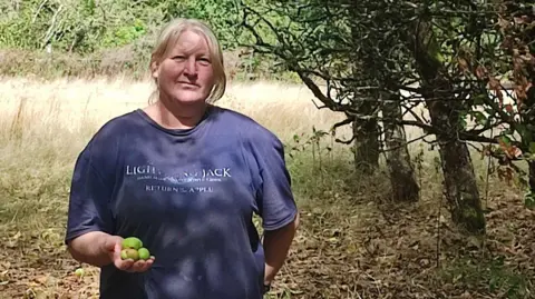Alison McIlwraith is standing in an orchard holding a handful of small green apples. Her blonde hair is tied back and she is wearing a blue T-shirt. The land around her looks very dry.