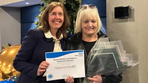 A woman with a navy blue suit poses with a woman wearing a black leather jacket and blonde hair. They both hold a certificate and the woman with blonde hair holds a bouquet of flowers.