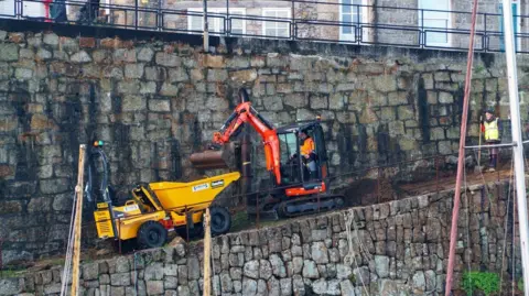 Historic England A small digger parked on a slope. The bucket of the digger is over a larger bucket. A man in a high vis jacket stands behind the digger and there is also one operating the machine.