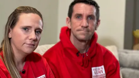 Lucy has long blonde hair and Mike has dark hair. They are both wearing red hoodies with Ollie's Army written on them. 