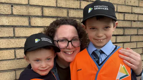 BBC Two young boys stand with their mother who is crouching between them and has her arms wrapped around them. They are smiling at the camera. The boy on the left wears black cap. Their mum has curly dark hair and glasses. The boy on the right wears a black cap, blue shirt, blue and grey tie, and an orange hi-vis jacket. 