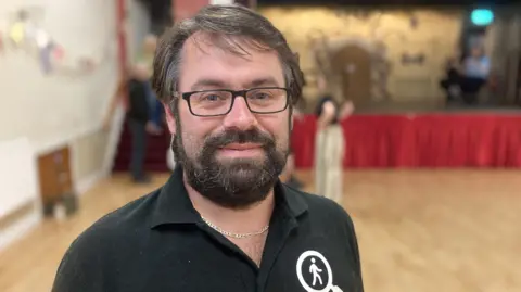 Adam Elms standing in front of a stage wearing a black polo shirt, back framed glasses with a grey and brown beard and hair.