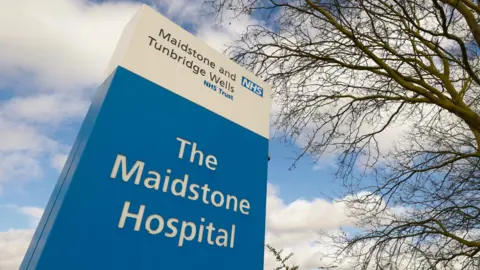 A large blue and white sign reading 'The Maidstone Hospital'. There is a blue and cloudy sky in the background.