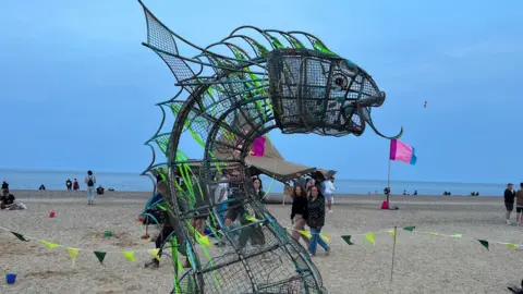 Martha Harrison/BBC A sculpture of an eel on Lowestoft beach. A group of young people walk past and look at it.