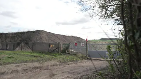 A general view of the waste storage site - metal gates and fences in front of a large mound of dirt and soil. 