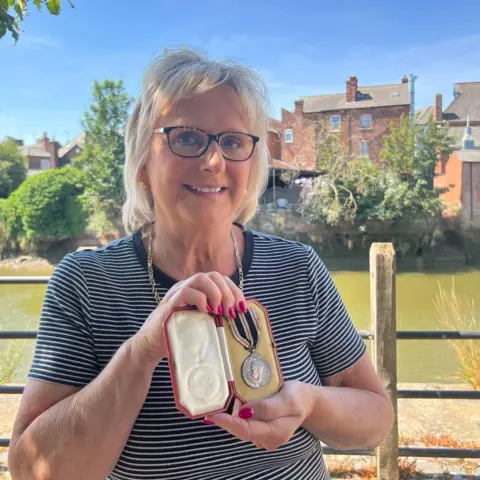 A woman wearing a stripy black and white top holding a silver medal with black and white ribbon housed in a red case with white and mustard-yellow inserts. She has blond hair and is standing in front of a river bank.