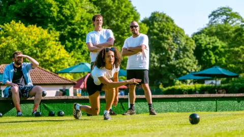 St George's Bowls Club A young woman lunges forward having thrown a bowls ball on grass, with three men behind her looking in its direction.