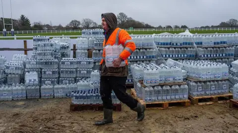 A person in an orange high vis jacket walking with water bottles under his arm. There are hundreds of bottles behind him.