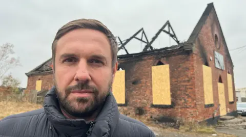 Adam Smith has a beard and brown hair. He is standing in front of the burnt-out gym which has a charred wooden roof and exposed beams.