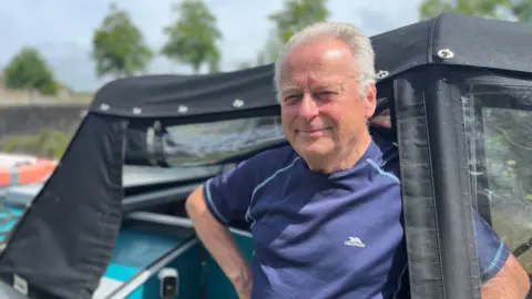A man with grey hair wearing a blue T-shirt stands with a black boat tarpaulin behind him. His boat is turquoise.