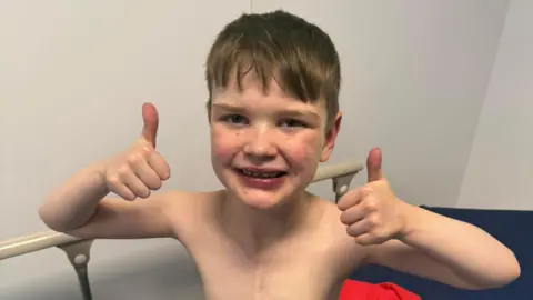 Máirtín Mac Gabhann A young boy sitting on a hospital bed smiling with both thumbs up.
