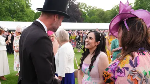 Prince William, who is wearing a suit and black top hat, speaks to a woman with wavy brown hair who is wearing a pink floral dress in a crowded garden area with a large white gazebo in the background.