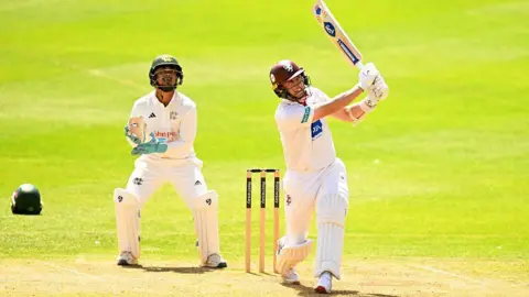 Getty Images Two people in cricket kit are on a green field. One of them is swinging a cricket bat. They are both looking in the direction of a ball which is out of shot.