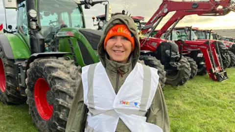 Lesely is wearing a green coat with a white reflective hi-viz jacket on top. She has an orange woolly hat on with Willdoes written on the front of it. She is standing in front of a line of tractors decorated with Christmas lights and decorations.