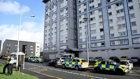 A block of flats clad in blue and white. Police cars line the residential streets and there is police tape blocking off the area, tied to a lamppost. Two police officers stand on the pavement.